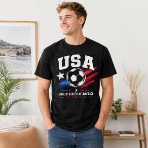 Puede incluir: Camiseta negra con un dise&ntilde;o gr&aacute;fico de bal&oacute;n de f&uacute;tbol. El dise&ntilde;o incluye la palabra "USA" en letras blancas, una estrella y pinceladas rojas y azules. Debajo est&aacute; el texto "UNITED STATES OF AMERICA SOCCER CLUB".