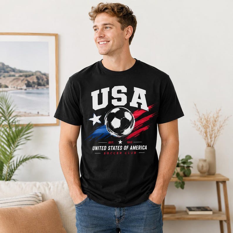 Puede incluir: Camiseta negra con un dise&ntilde;o gr&aacute;fico de bal&oacute;n de f&uacute;tbol. El dise&ntilde;o incluye la palabra "USA" en letras blancas, una estrella y pinceladas rojas y azules. Debajo est&aacute; el texto "UNITED STATES OF AMERICA SOCCER CLUB".