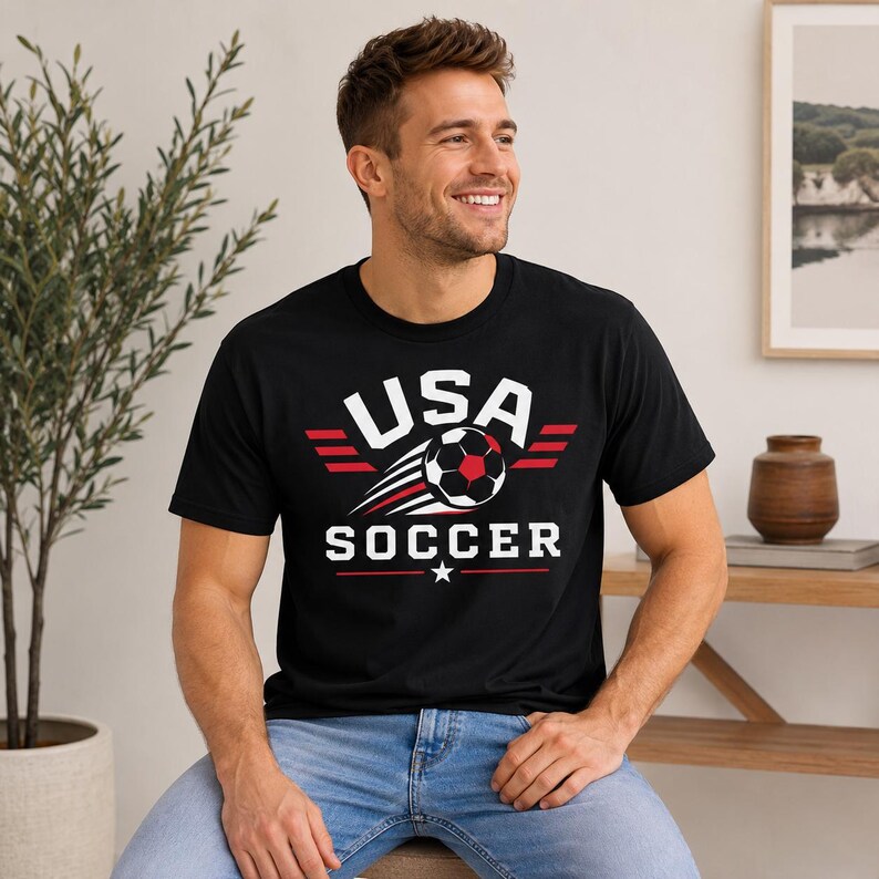 May include: Black t-shirt featuring the text "USA SOCCER" in white, red, and black. The design includes a soccer ball and wing-like stripes. The shirt is made of a soft material.