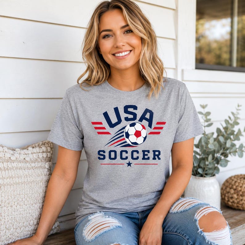 May include: A grey t-shirt with the text "USA SOCCER" in navy blue and red, featuring a soccer ball graphic. The shirt has a crew neck and short sleeves. The design includes a star and wing-like elements.