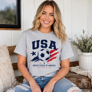 Puede incluir: Camiseta gris con un dise&ntilde;o de bal&oacute;n de f&uacute;tbol y la palabra "USA" en azul marino. El dise&ntilde;o incluye una estrella, pinceladas rojas y azules, y el texto "United States of America Soccer Club."