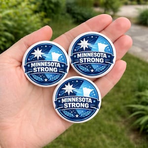 May include: Three round, white and blue buttons with the text "MINNESOTA STRONG". Each button features a star, a map outline, and a banner. The buttons are held in a hand against a blurred green background.