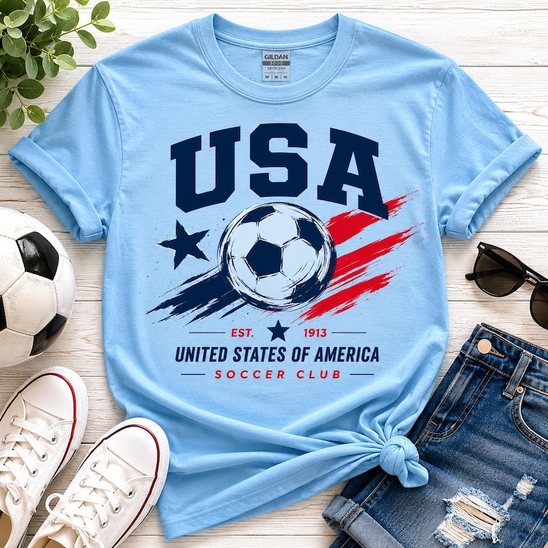 Puede incluir: Camiseta azul claro con un dise&ntilde;o "USA" en azul marino, un bal&oacute;n de f&uacute;tbol y el texto "United States of America Soccer Club". El dise&ntilde;o incluye una estrella y pinceladas rojas y azules. Se ve un bal&oacute;n de f&uacute;tbol y zapatillas blancas.