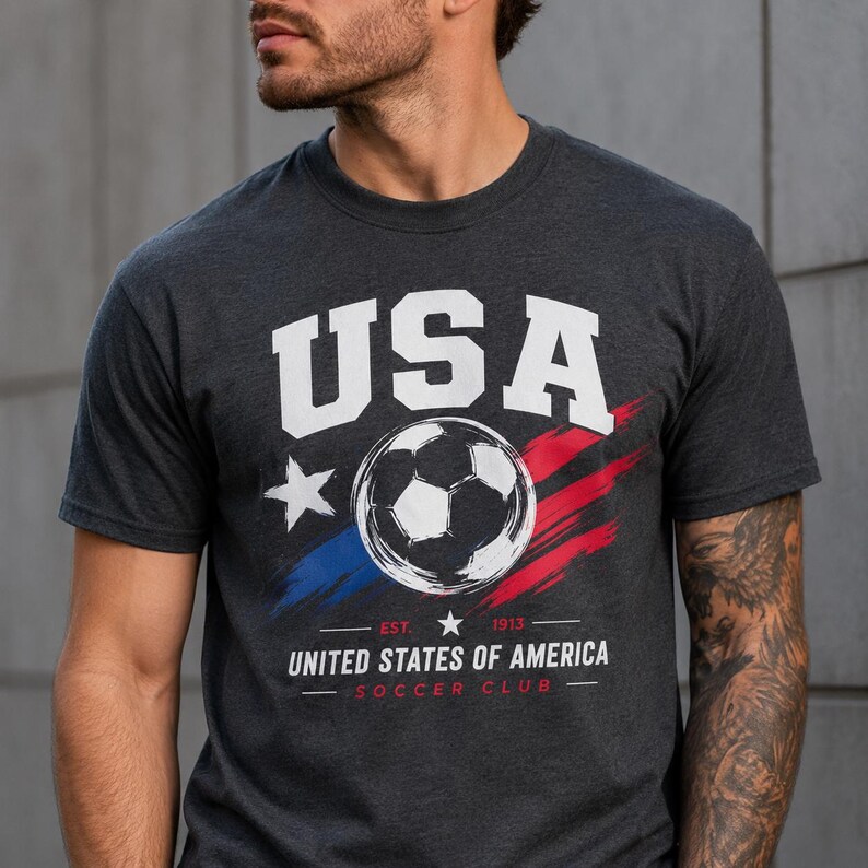 Puede incluir: Camiseta gris oscuro con un gr&aacute;fico de bal&oacute;n de f&uacute;tbol. La camiseta presenta la palabra "USA" en letras blancas en bloque sobre un bal&oacute;n de f&uacute;tbol y pinceladas rojas y azules. Debajo, el texto "UNITED STATES OF AMERICA SOCCER CLUB".