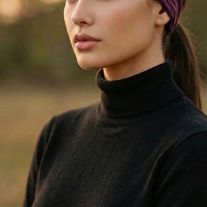 Puede incluir: Una persona con un jersey de cuello alto negro y una diadema morada oscura. La diadema es ancha y cubre la frente. El fondo est&aacute; borroso, con una luz natural suave.