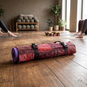 May include: Rolled purple yoga mat secured with black straps inside a patterned red and burgundy carrying case. The case has a handle and buckle closures. The mat is on a wooden floor in a yoga studio.