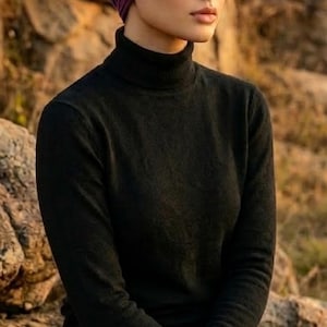 Puede incluir: Una mujer con un turbante de punto acanalado morado oscuro y un jersey de cuello alto negro. La imagen es un plano medio, con el rostro y la parte superior del cuerpo de la mujer enfocados. El fondo est&aacute; borroso, con luz natural.