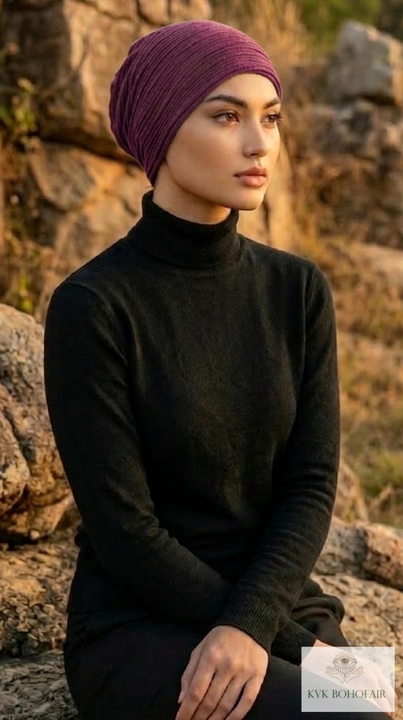 Puede incluir: Una mujer con un turbante de punto acanalado morado oscuro y un jersey de cuello alto negro. La imagen es un plano medio, con el rostro y la parte superior del cuerpo de la mujer enfocados. El fondo est&aacute; borroso, con luz natural.