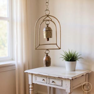 May include: A hanging metal wind chime featuring a gold-toned arched frame with two weathered, rustic bells suspended from the centre and bottom. The structure is connected by a circular ring at the top, showcasing a metallic finish throughout.