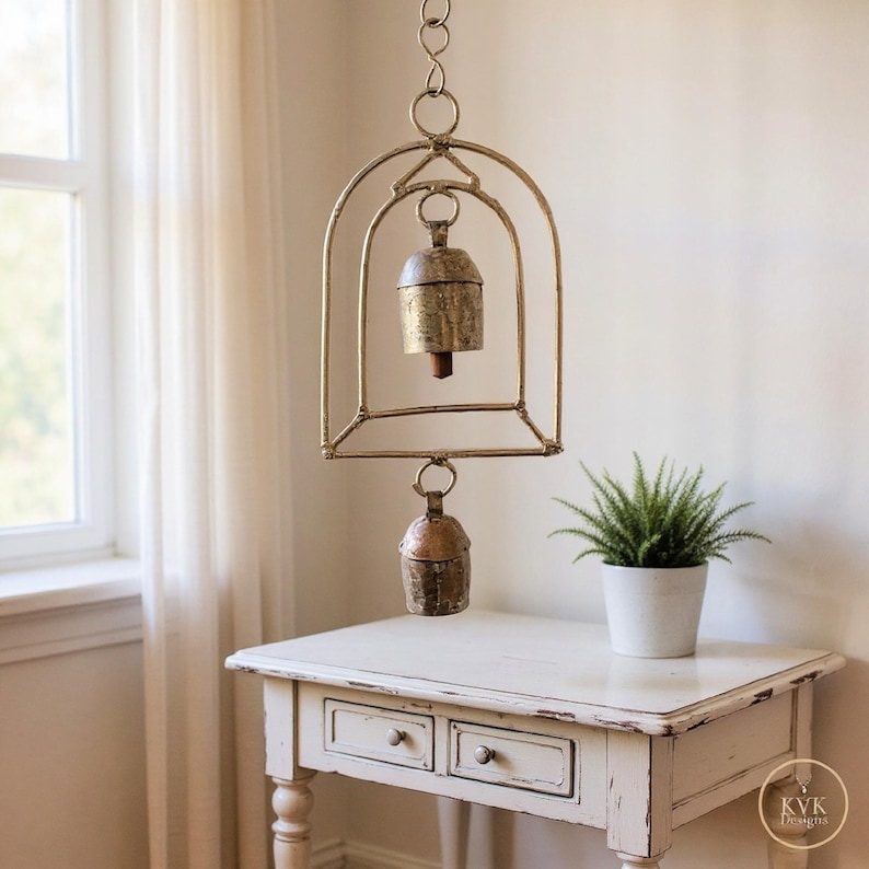 May include: A hanging metal wind chime featuring a gold-toned arched frame with two weathered, rustic bells suspended from the centre and bottom. The structure is connected by a circular ring at the top, showcasing a metallic finish throughout.