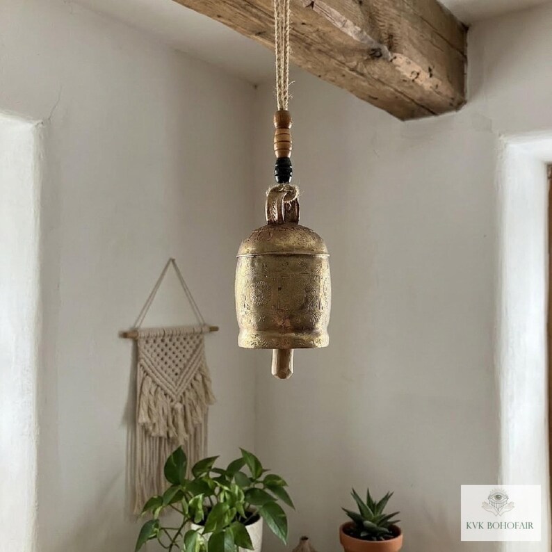 May include: A hanging brass bell with a weathered, textured finish suspended by a jute rope. The rope features a small wooden bead and a black bead above the bell. The bell has a cylindrical body with a flared base and a central clapper.