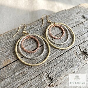 May include: A pair of dangle earrings featuring three interlocking circular rings in gold, silver, and copper tones. Each ring has a textured, hammered finish. The earrings are suspended from gold-toned ear wires.