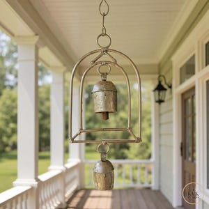 May include: A hanging metal bell wind chime with two bells. The bells are a tarnished gold colour and are suspended within a decorative metal frame. The frame has a curved top and a rectangular base. The chime is hanging on a porch.