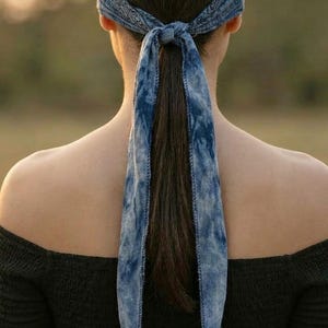 May include: A blue tie-dye bandana tied around the head and into a ponytail. The bandana is a light blue with a darker blue tie-dye pattern. The person is wearing a black off-the-shoulder top.