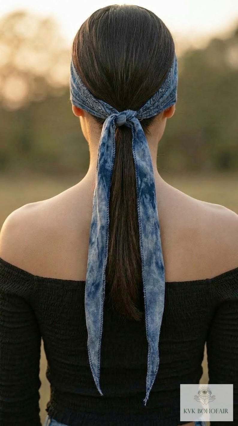May include: A blue tie-dye bandana tied around the head and into a ponytail. The bandana is a light blue with a darker blue tie-dye pattern. The person is wearing a black off-the-shoulder top.
