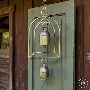 May include: A hanging metal wind chime featuring a curved arch frame with two silver-toned bells suspended vertically. The upper bell is housed within the arch, while the lower bell hangs below. The metal has a weathered finish and includes a small logo tag reading KVK Designs.