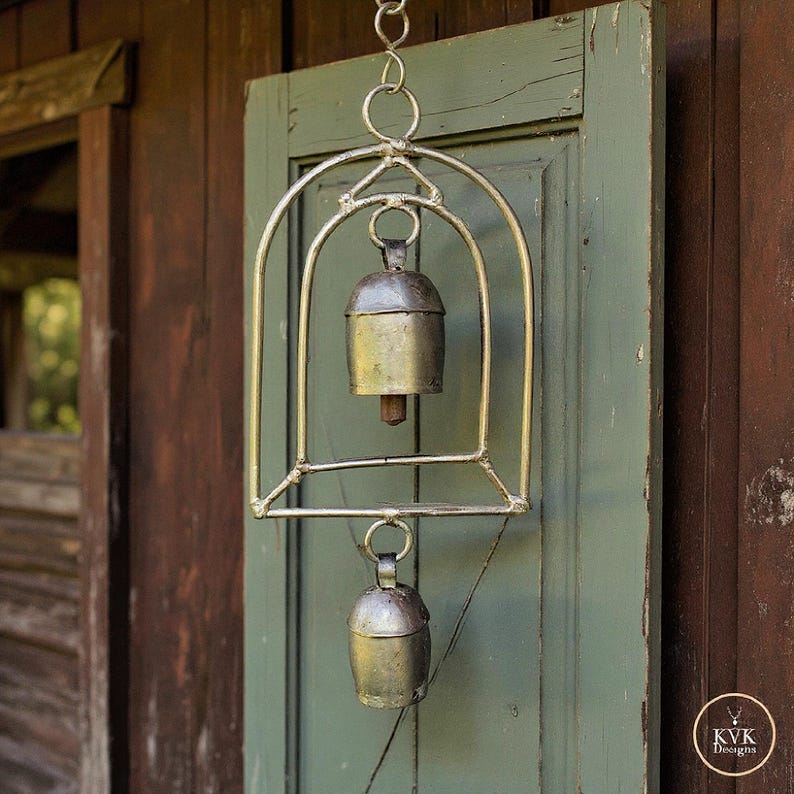 May include: A hanging metal wind chime featuring a curved arch frame with two silver-toned bells suspended vertically. The upper bell is housed within the arch, while the lower bell hangs below. The metal has a weathered finish and includes a small logo tag reading KVK Designs.