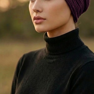 Puede incluir: Primer plano de una mujer con un gorro de punto acanalado morado oscuro y un jersey de cuello alto negro. La imagen se centra en la parte superior del cuerpo y la cara. El fondo est&aacute; borroso, con una paleta de colores c&aacute;lidos y naturales.