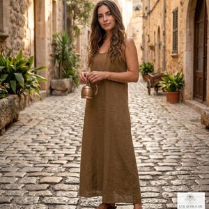 May include: A floor-length, sleeveless brown dress with a simple scoop neckline and a relaxed, straight-cut silhouette. The garment features a plain, solid-colored fabric and is worn with brown sandals. A small, metallic bell is held in hand.