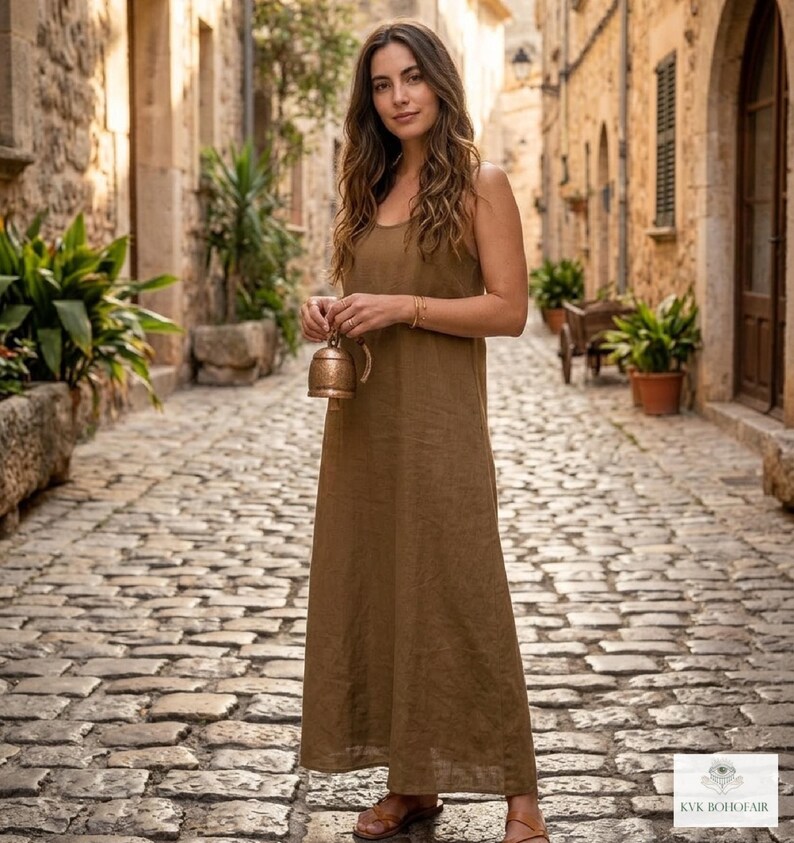 May include: A floor-length, sleeveless brown dress with a simple scoop neckline and a relaxed, straight-cut silhouette. The garment features a plain, solid-colored fabric and is worn with brown sandals. A small, metallic bell is held in hand.