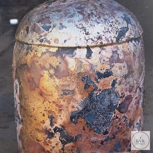 May include: A cylindrical container with a lid, featuring a heavily textured, weathered surface in shades of metallic gold, copper, and dark charcoal black. The lower right corner displays a small logo with the text KVK Designs.
