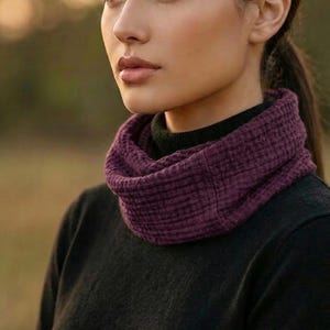 Puede incluir: Una mujer con un cuello de bufanda texturizado de color p&uacute;rpura oscuro. La bufanda se combina con un su&eacute;ter negro. El fondo est&aacute; borroso, con una paleta de colores c&aacute;lidos y naturales. El logotipo "KVK BOHOFAIR" es visible.