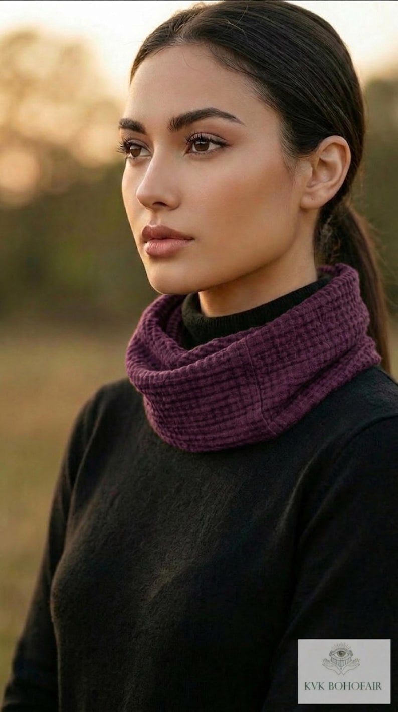 Puede incluir: Una mujer con un cuello de bufanda texturizado de color p&uacute;rpura oscuro. La bufanda se combina con un su&eacute;ter negro. El fondo est&aacute; borroso, con una paleta de colores c&aacute;lidos y naturales. El logotipo "KVK BOHOFAIR" es visible.