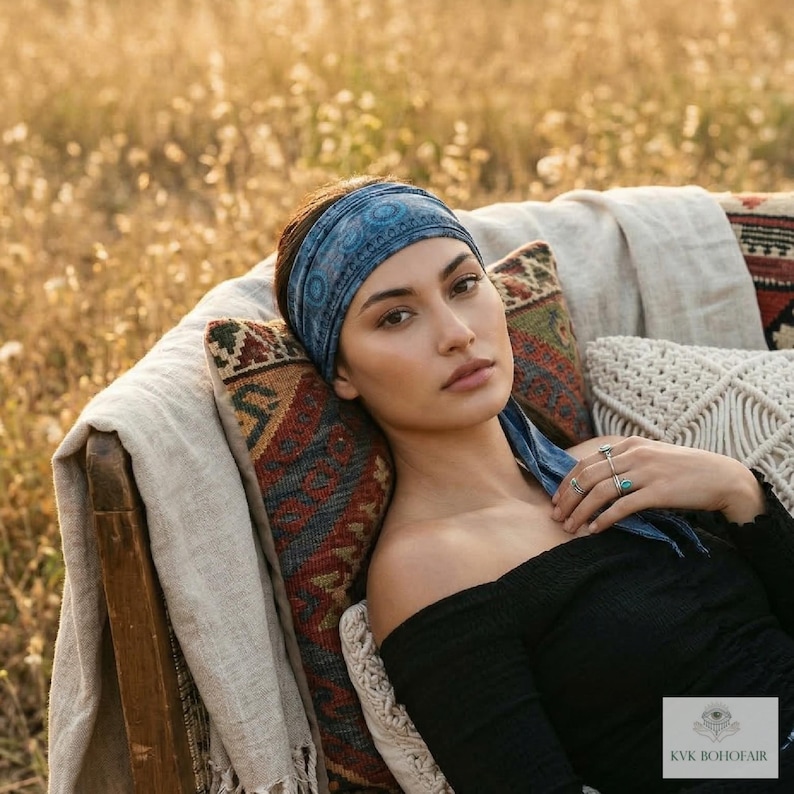 May include: A woman wearing a blue patterned headband and black off-the-shoulder top. She is reclining on a wooden chair with patterned pillows and a cream-colored blanket. The background is a field of tall grass.