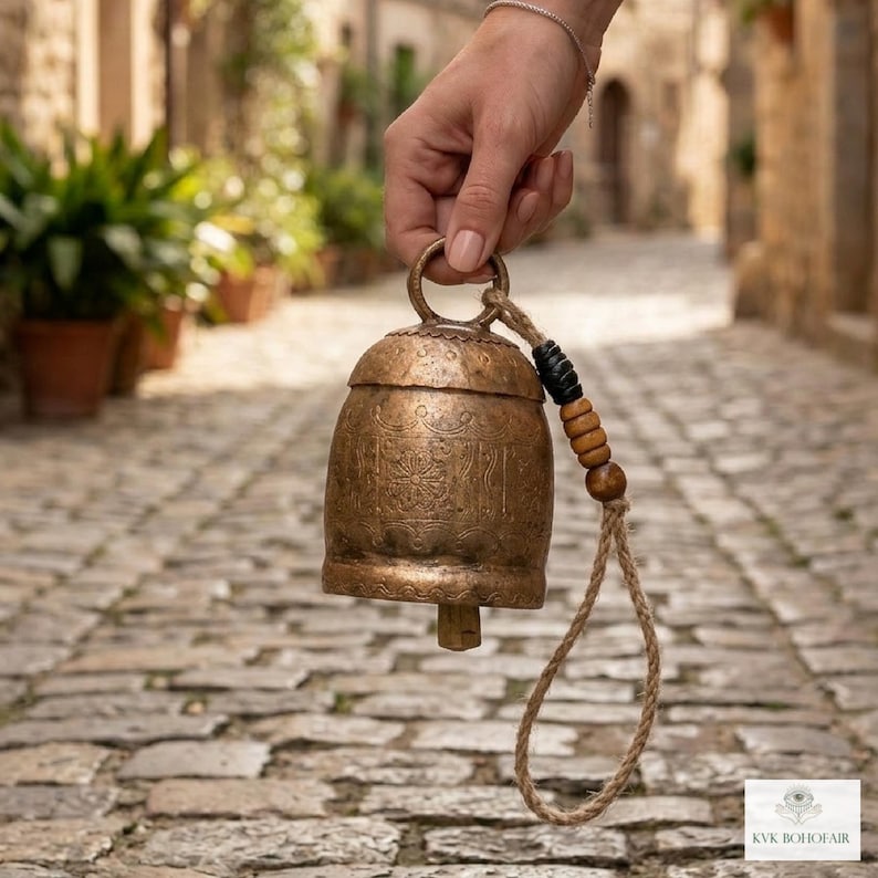 May include: A weathered bronze-colored metal bell featuring intricate etched patterns on its surface. The bell includes a sturdy loop handle attached to a thick jute rope adorned with one black and three wooden beads for a textured appearance.