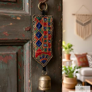 May include: Embroidered wall hanging with a brass bell. The rectangular textile features a geometric pattern in red, blue, green, and gold thread. It hangs from a black strap and a brass ring, attached to a weathered wooden door.
