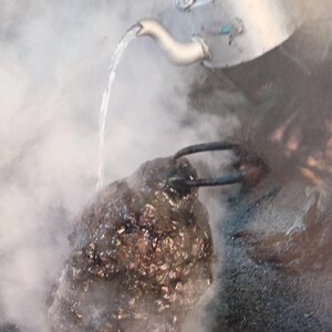 May include: A silver-colored metal kettle pours a stream of water onto a dark, textured, irregular-shaped stone object. The scene is filled with thick white steam rising from the point of contact. A small logo with the text KVK Designs is visible.