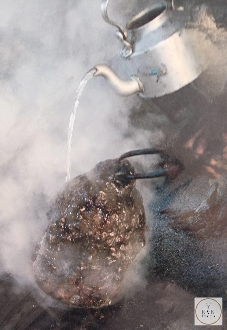 May include: A silver-colored metal kettle pours a stream of water onto a dark, textured, irregular-shaped stone object. The scene is filled with thick white steam rising from the point of contact. A small logo with the text KVK Designs is visible.