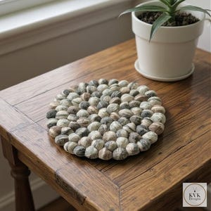 Puede incluir: Un salvamanteles redondo hecho de pequeñas bolas de lana afieltrada en tonos grises, marrones y crema. El salvamanteles está colocado sobre una mesa de madera junto a una planta en maceta blanca. El salvamanteles tiene aproximadamente 25 cm de diámetro.