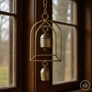May include: A decorative hanging bell made of tarnished gold metal. The bell is suspended within a cage-like frame, hanging from a chain. The frame has an arched design. The background is a dark wooden window frame.
