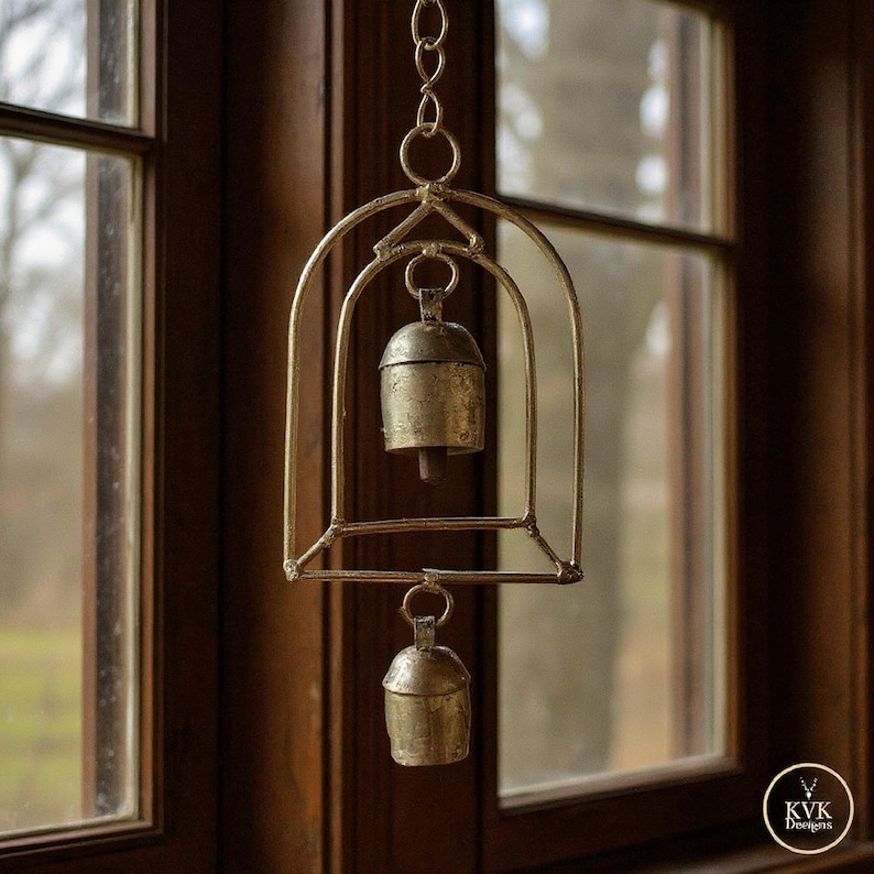 May include: A decorative hanging bell made of tarnished gold metal. The bell is suspended within a cage-like frame, hanging from a chain. The frame has an arched design. The background is a dark wooden window frame.
