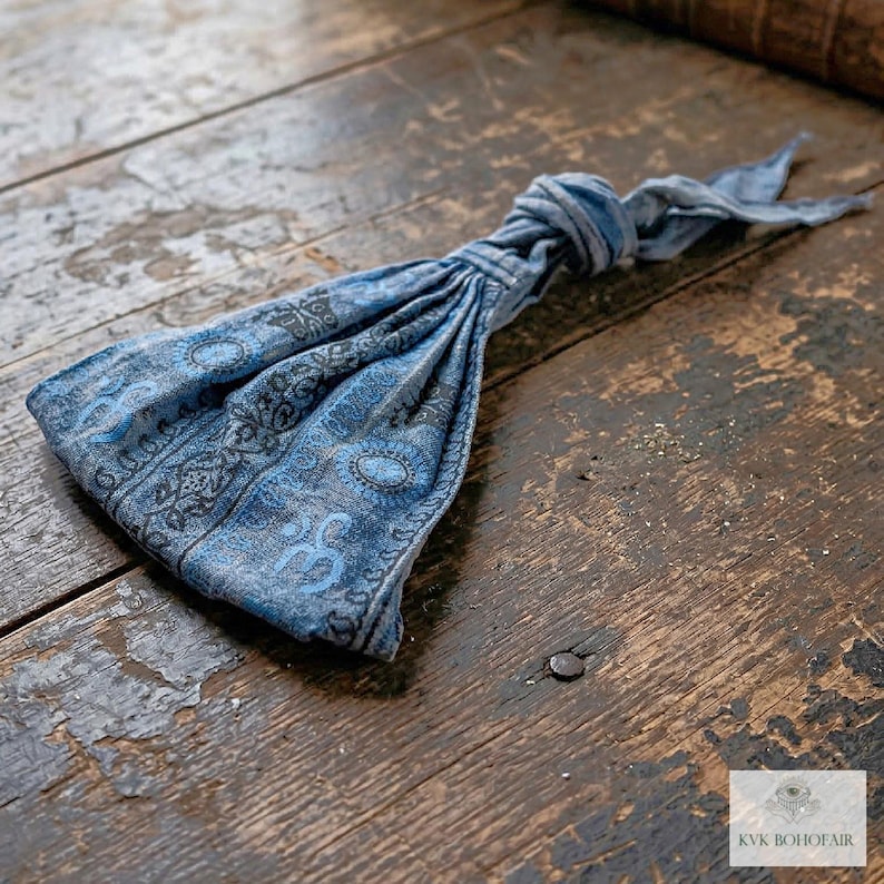 May include: A blue patterned headband with a knotted top, resting on a rustic wooden surface. The headband features a detailed design in shades of blue and gray. The fabric appears soft and flexible, suitable for various head sizes.