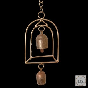 May include: A handcrafted wind chime featuring two small, aged metal bells suspended within a gold-coloured geometric frame. The bells have a rustic, antique appearance, and the frame is connected by a chain. The design includes the text "KVK Designs".