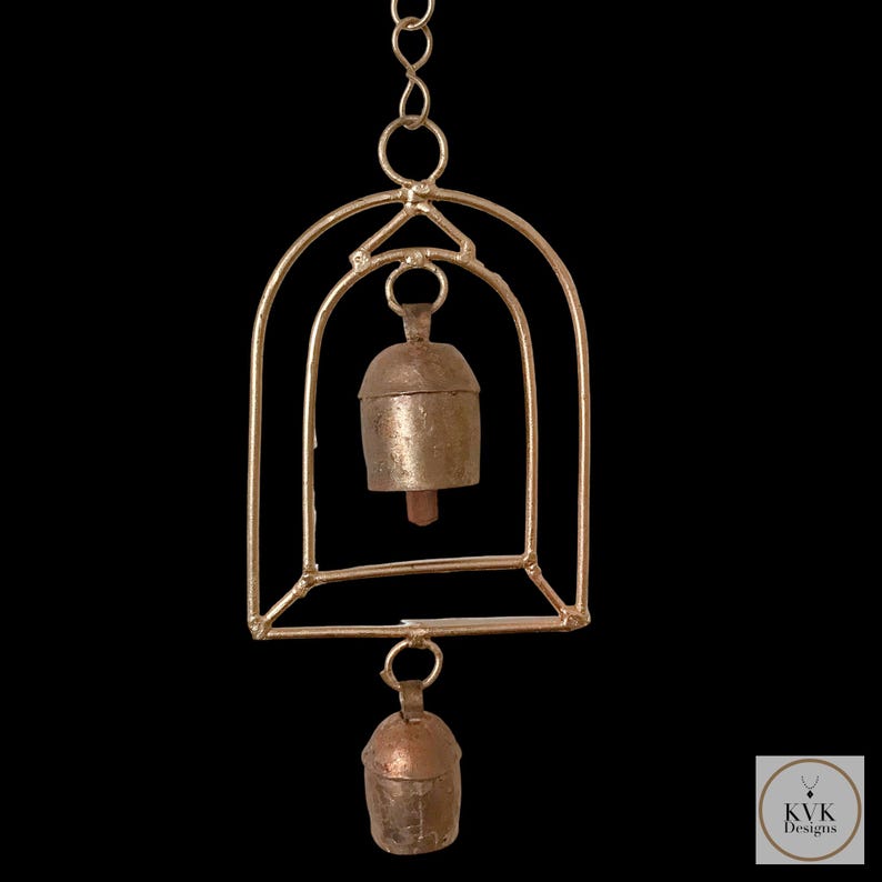 May include: A handcrafted wind chime featuring two small, aged metal bells suspended within a gold-coloured geometric frame. The bells have a rustic, antique appearance, and the frame is connected by a chain. The design includes the text "KVK Designs".
