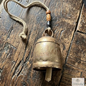 May include: A brass bell with etched floral and geometric patterns, featuring a wooden clapper inside. The bell hangs from a jute rope adorned with two light brown wooden beads and one dark, textured bead above the metal loop.