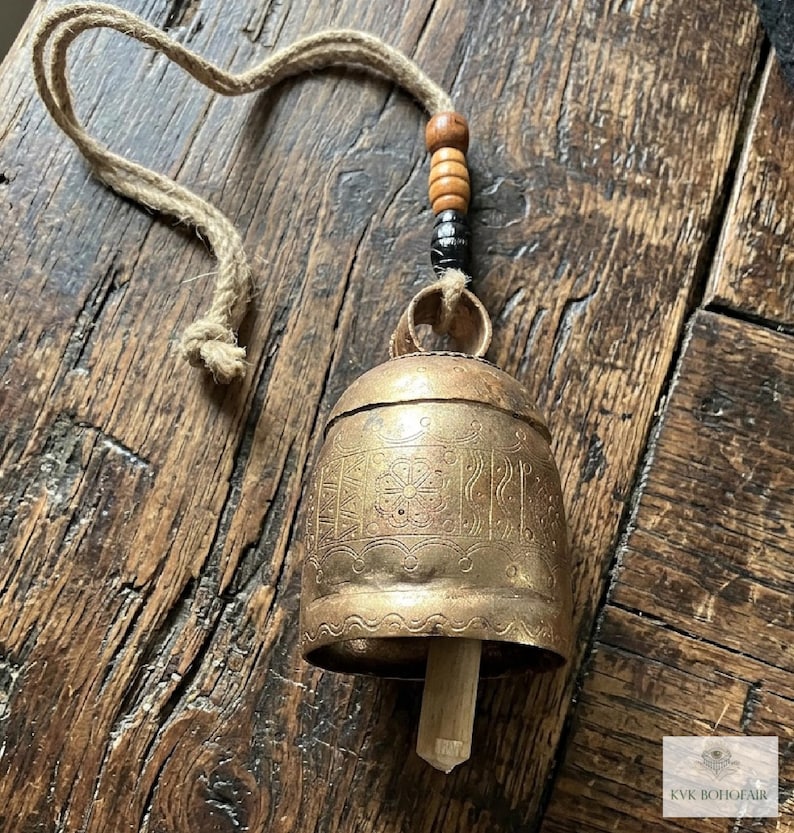 May include: A brass bell with etched floral and geometric patterns, featuring a wooden clapper inside. The bell hangs from a jute rope adorned with two light brown wooden beads and one dark, textured bead above the metal loop.