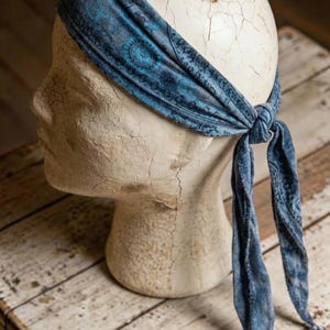 May include: A blue paisley patterned headband tied at the back, displayed on a mannequin head. The headband is a soft, textured fabric with a dark blue and teal design. The mannequin head is off-white and sits on a rustic wooden surface.