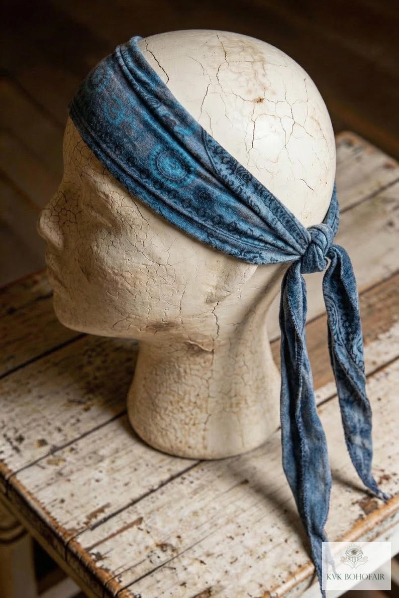 May include: A blue paisley patterned headband tied at the back, displayed on a mannequin head. The headband is a soft, textured fabric with a dark blue and teal design. The mannequin head is off-white and sits on a rustic wooden surface.