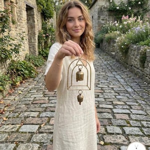 May include: A brass bell chime with an arched frame is held up. The chime has two bells, one larger than the other. The person holding the chime is wearing a cream-coloured dress. The background is a cobbled street.