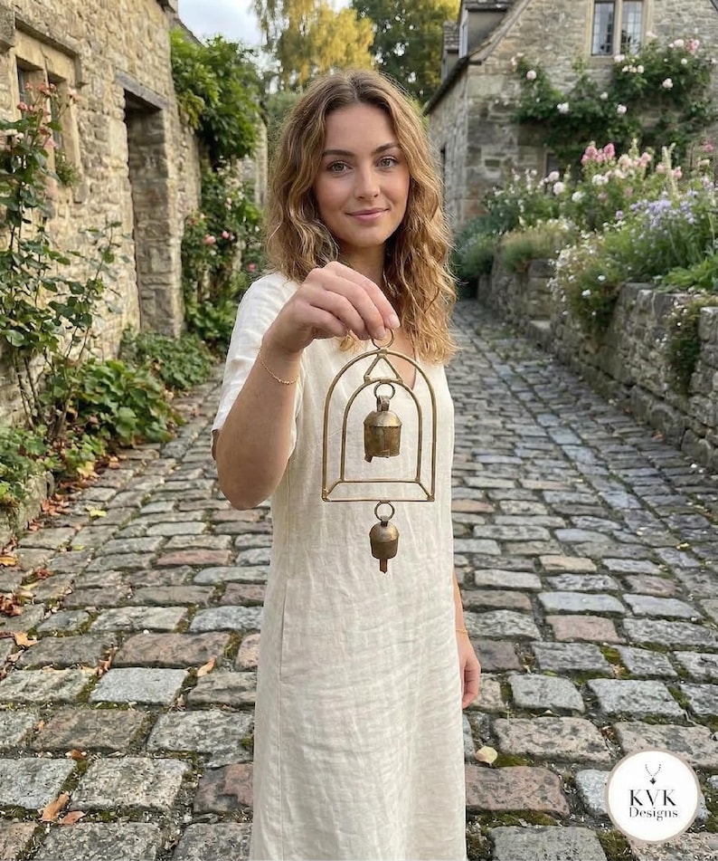 May include: A brass bell chime with an arched frame is held up. The chime has two bells, one larger than the other. The person holding the chime is wearing a cream-coloured dress. The background is a cobbled street.