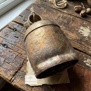 May include: A weathered brass bell featuring intricate etched floral and geometric patterns, including a central zig-zag design. The bell has a rustic, tarnished finish and a small loop at the top with a piece of twine. A small tag reads KVK BOHOFAIR.