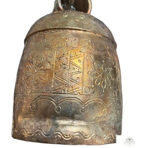 May include: A metallic bell with a weathered gold finish, featuring embossed floral patterns and geometric line designs. The bell has a rounded top loop and a scalloped border near the base. The text KVK BOHOFAIR is visible at the bottom.