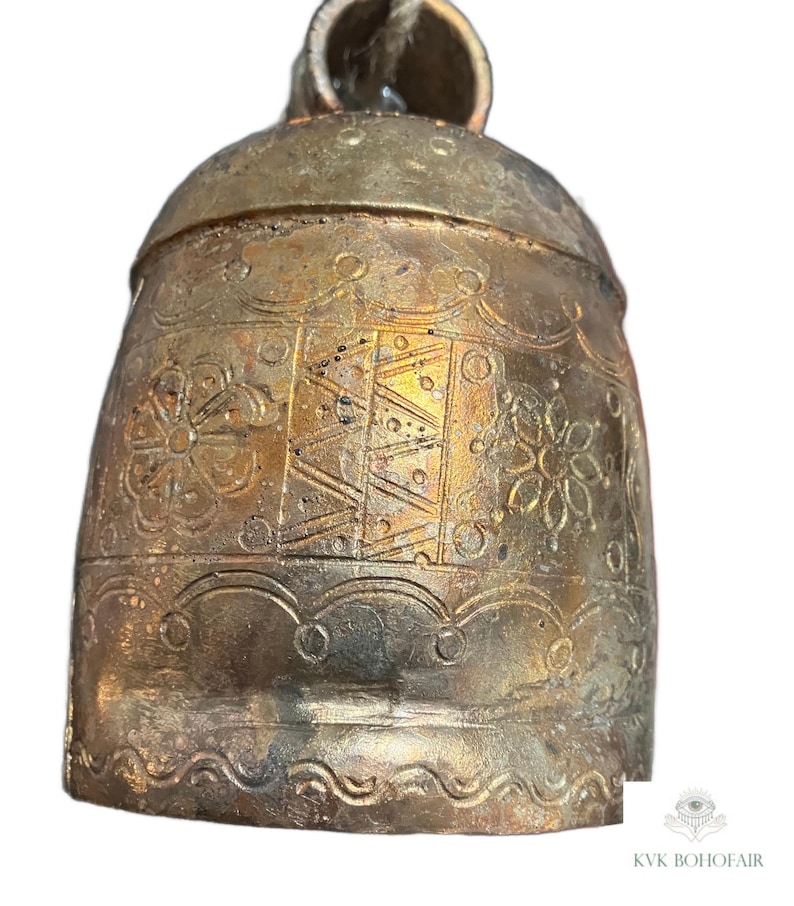 May include: A metallic bell with a weathered gold finish, featuring embossed floral patterns and geometric line designs. The bell has a rounded top loop and a scalloped border near the base. The text KVK BOHOFAIR is visible at the bottom.