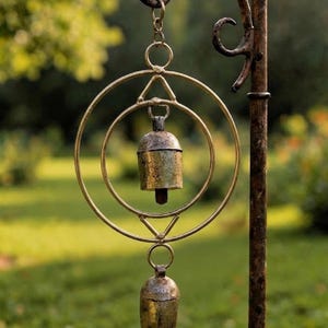 May include: A rustic metal wind chime with two bells suspended within a circular and triangular frame. The chime hangs from a curved metal arm attached to a vertical pole. The bells and frame have a weathered, antique finish.