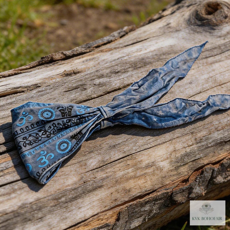 May include: A blue and gray patterned headband with a tied bow design. The headband features a repeating pattern of the Om symbol and other decorative elements. The headband is made of a soft, textured fabric and is displayed on a weathered wooden surface.