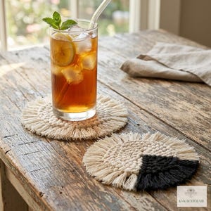 May include: A tall glass of iced tea with lemon and mint sits on a cream-colored macrame coaster. A second coaster with a black accent is next to it. The coasters are on a rustic wooden table.